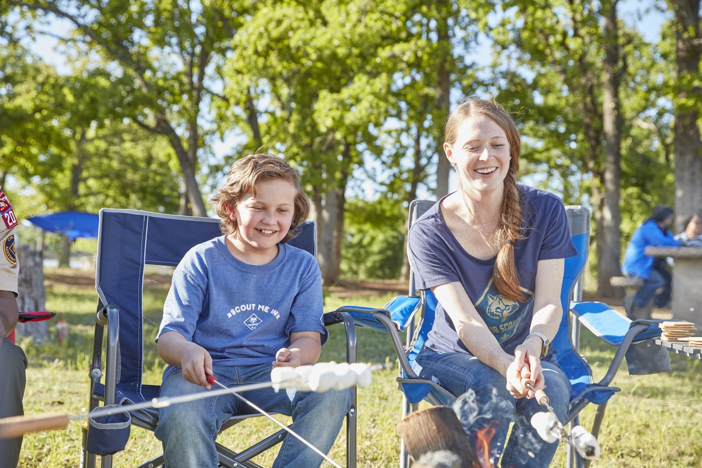 A Cub Scout and a parent toasting marshmallows over a campfire in camping chairs.
