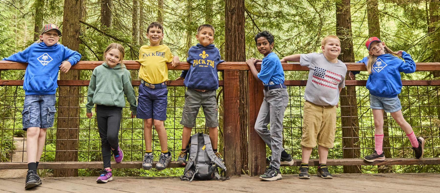 Seven Cub Scouts lined up along a wooden fence rail in a redwood forest, grinning at the camera.